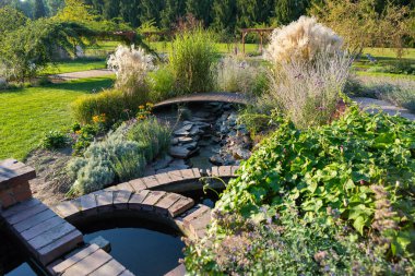 Sakin bir bahçe, renkli çiçekler ve yeşilliklerle sarılmış dolambaçlı bir gölet sergiliyor. Canlı bitkiler açık mavi gökyüzünün altında gelişerek, barışçıl bir geri çekilme yaratırlar..