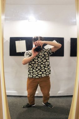 Aşırı kısa bacaklı, çiçekli bir gömlek giyen genç bir adam optik illüzyon müzesinde fotoğraf çekiyor. Adam aynaya yansıyor..