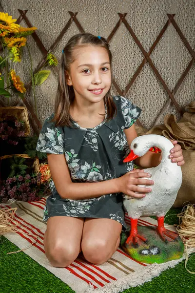 Çiçekli elbiseli, at kuyruklu, elinde bir örtü, elinde kaz olan bir kız. Saman ve ahşap çitli taşralı bir arka plan. İkisi de kırsal kesimde kameraya bakıyor..