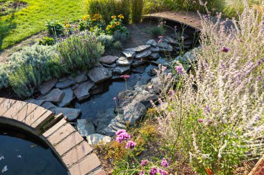 Resimli bir bahçe sahnesinde yemyeşil ve renkli çiçeklerle çevrili hafif akan bir dere sergileniyor. Huzurlu atmosfer rahatlamayı ve doğayla bağlantıyı davet eder..