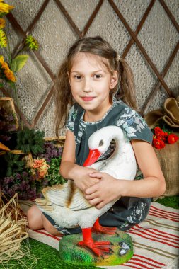 Uzun kahverengi saçlı ve çiçekli elbiseli bir kız yerde beton bir kazın yanında oturuyor. Kız gülümsüyor ve kaza sarılıyor. Arka planda çiçekler ve sepetler var..