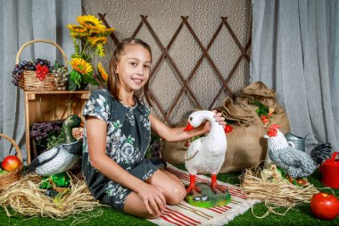Genç bir kız bir ördekle ahırda diz çöküyor. Onun yanında bir sepet sebze ve meyve var. Arka planda ahşap çitler ve saman balyası var..