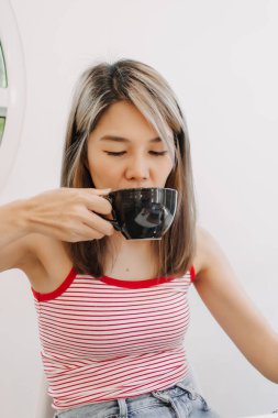 Kırmızılı tatlı Asyalı kız, siyah fincanda sıcak kahve içiyor..