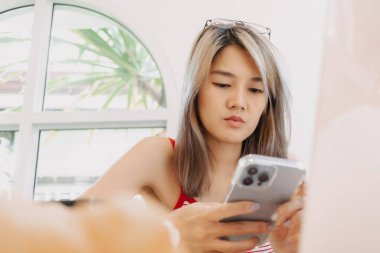 Şirin serbest çalışan Asyalı kız dizüstü bilgisayar üzerinde çalışırken akıllı telefon kullanıyor..