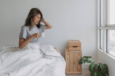 Asyalı kadın beyaz yatak odasında boş bir fincan kahve tutuyor..