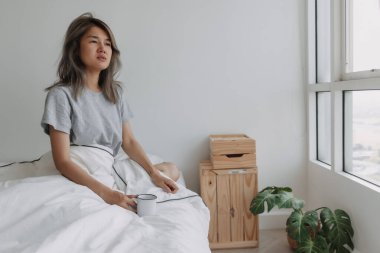 Asyalı kadın beyaz yatak odasında boş bir fincan kahve tutuyor..