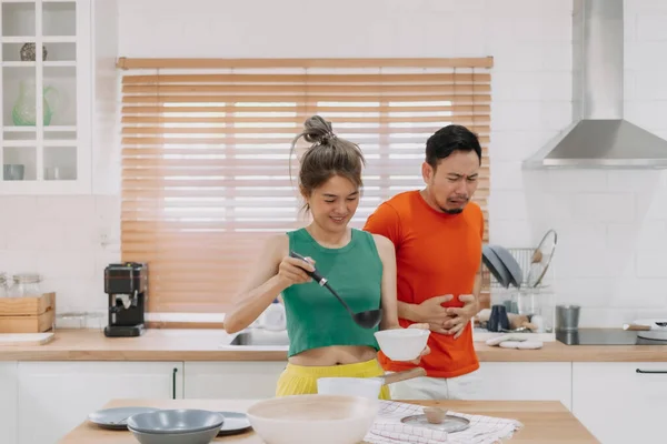 Asian couple. Funny shocked face reaction of husband looking at wife cooking.