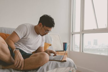 Asyalı bir adam odasında bilgisayarla sıkıcı bir işi uyandırıyor. Kanepede oturuyor.. 