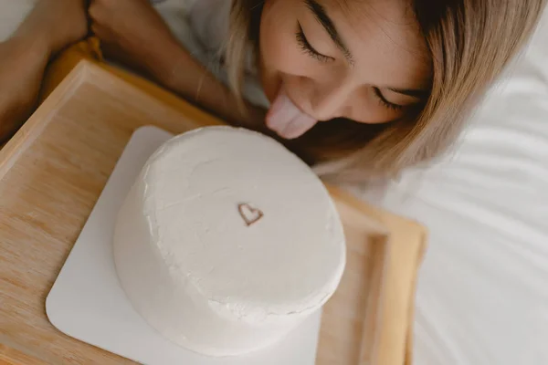 Close up of funny Asian woman tongue out trying to eat white minimal heart cake on the bed.