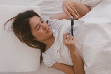 Asyalı kadın cep telefonuyla sosyal medya yayınlarını kontrol ediyor, uyuyamıyor ve uyanık kalamıyor.