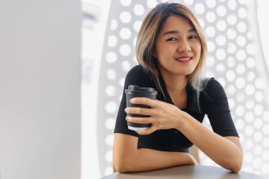 Siyah tişörtlü, beyaz kahve dükkanında sıcak kahve yudumlayan mutlu Asyalı kadın..