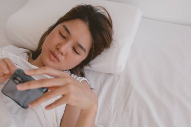 Asyalı kadın cep telefonuyla sosyal medya yayınlarını kontrol ediyor, uyuyamıyor ve uyanık kalamıyor.