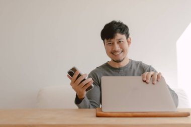 Bilgisayarda çalışan mutlu Asyalı adam cep telefonundaki uygulamadan memnun..