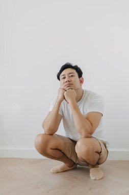 Sıkıcı suratlı Asyalı adam beyaz duvarlı yerde çömelmiş..