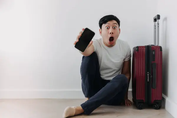 Shock and surprise face of asian man using mobile phone application for travel, sitting with his baggage or luggage.