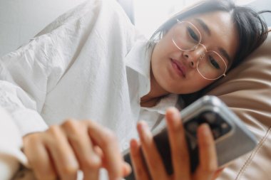 Asyalı Taylandlı bir kadının kahverengi kanepeye uzanıp dinlenirken cep telefonu kullanırken gözlük takması işten bıkmış.. 