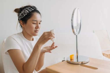 Asyalı Taylandlı kadın aynanın önünde beyaz koltukta otururken güneş kremi kullanıyor ve uyguluyor.. 