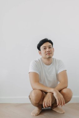 Sıkıcı suratlı Asyalı adam beyaz duvarlı yerde çömelmiş..