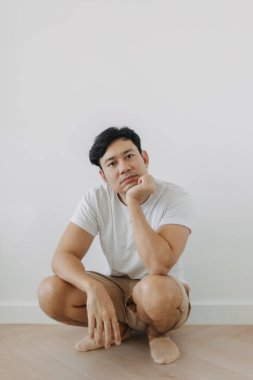 Sıkıcı suratlı Asyalı adam beyaz duvarlı yerde çömelmiş..