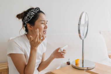 Asyalı Taylandlı kadın aynanın önünde beyaz koltukta otururken güneş kremi kullanıyor ve uyguluyor.. 