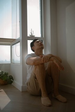 Üzgün ve yalnız Asyalı adam yerde oturmuş dizine sarılıyor..