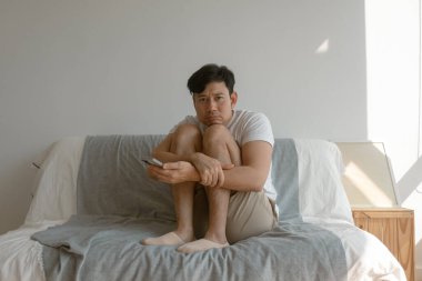 Yalnız Asyalı bir adam dizlerine sarılıyor koltukta oturuyor ve cep telefonundan sosyal medyayı kontrol ediyor..