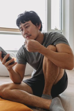 Asyalı bir adam yerde oturmuş çerez yiyor ve cep telefonuna bakıyor.