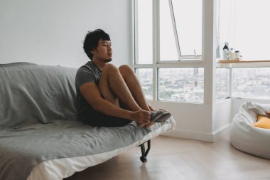 Gri tişörtlü Asyalı bir adam evinin oturma odasında çorap giyiyor..