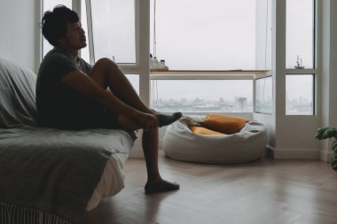 Gri tişörtlü Asyalı bir adam evinin oturma odasında çorap giyiyor..