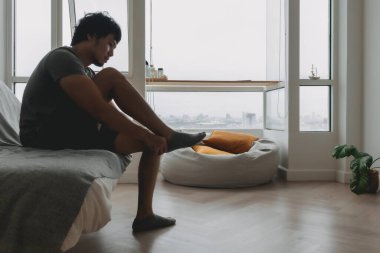 Gri tişörtlü Asyalı bir adam evinin oturma odasında çorap giyiyor..