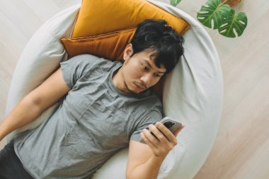 Asyalı adam oturma odasındaki akıllı telefonu kullanarak kanepede uzanıyor..
