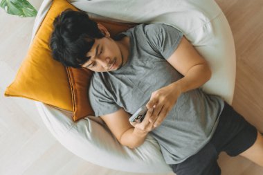 Asyalı adam oturma odasındaki akıllı telefonu kullanarak kanepede uzanıyor..