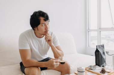 Boş kafalı Asyalı adam elinde kahve fincanıyla pencereden dışarı bakıyor..