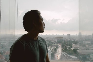 Yalnız, bekar Asyalı bir adam apartmanın penceresinden dışarı bakıyor..