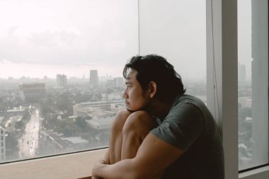 İçine kapanık Asyalı adam dizlerine sarılıp dışarı bakıyor..