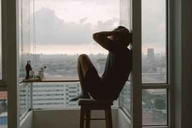 İçine kapanık yalnız Asyalı adam pencerelerde oturuyor. Evde yalnız olmanın tadını çıkarıyor..