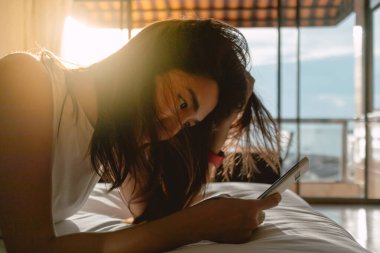 Asyalı kadın cep telefonuyla yatakta dinleniyor, sakinliğin ve sıcak güneşin tadını çıkarıyor..