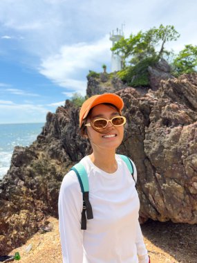 Arkasında okyanus ve gökyüzü olan kayaların yanında mutlu poz veren bir turist.