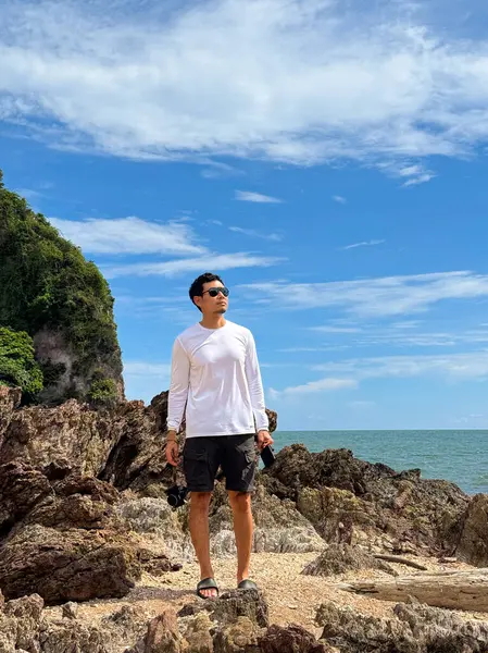 Beyaz tişörtlü, kendinden emin Asyalı erkek açık gökyüzünün altında kayalık kıyı şeridinde duruyor.