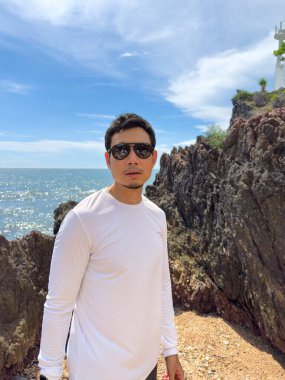 Kayaların yanında duran, arkasında okyanus ve bulutlu gökyüzü olan bir adam.