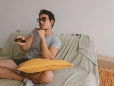 Genç Asyalı erkek kanepede bağdaş kurup uzaktan kumandayı tutuyor ve gün ışığında sakince televizyon izliyor..