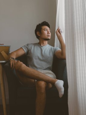 Rahat Taylandlı bir erkek, rahat bir Tayland iç mekanında, pencerenin yanındaki tahta bir sandalyede otururken kollarını uzatır ve rahat ve doğal vücut dilini gösterir..