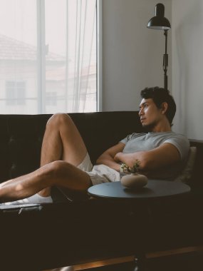 Rahatlamış Taylandlı erkek, elleri göğsünde, bacakları açık, koyu deri bir kanepede uzanmış, sakin ve durgun bir dairede dinleniyor..