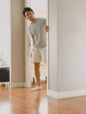 Taylandlı adam bej bir duvarın arkasından ahşap zeminli parlak bir daireye bakıyor, sakin bir ev ortamında neşeli bir enerji gösteriyor..