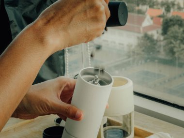 El değirmeni ve kupa kullanarak, arka planda bulanık şehir binaları ve sıcak güneş ışığıyla, Asyalı el yapımı kahve hazırlıyor.