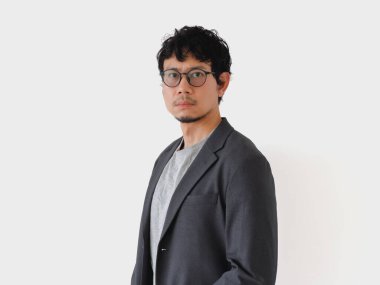 Takım elbiseli ve gözlüklü bir adam beyaz bir duvarın önünde duruyor. Ciddi ve odaklanmış görünüyor.