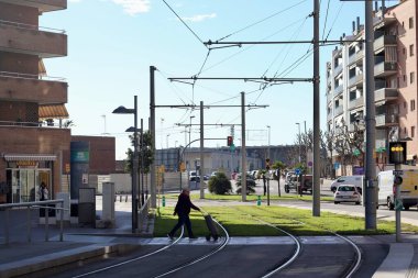 Sant Joan Despi, İspanya - 21 Aralık 2024: Güneşli bir günde modern binalar, sokak lambaları ve yeşil bantlarla çevrili kentsel bir mahallede tramvay ile tramvay raylarından geçen bir kişi