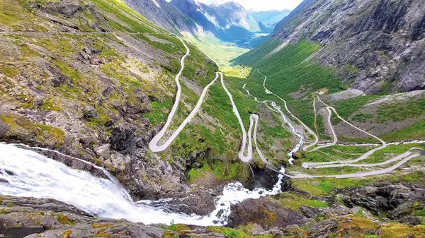 Norveç 'teki ünlü Trollstigen dağ yolunun havadan görünüşü, sarp kayalıklardan dökülen güçlü bir şelale. Serpentine yolu açık mavi gökyüzünün altında dramatik alp manzarası boyunca esiyor..