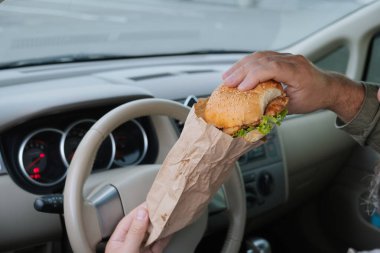 Arabada hamburger tutan bir adamın ellerinin yakın çekimi. Bir el kese kağıdındaki hamburgeri destekliyor, diğer el ekmeğin üstünü tutuyor. Yolda ya da otoparkta atıştırmalık bir şeyler..