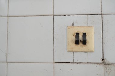 Beyaz kiremitlerle kaplı bir duvara eski bir çift klavyeli anahtar monte edilmiş. Anahtarın yüzeyinde ve fayanslarda zaman izleri var: toz, kir ve sürtünme..
